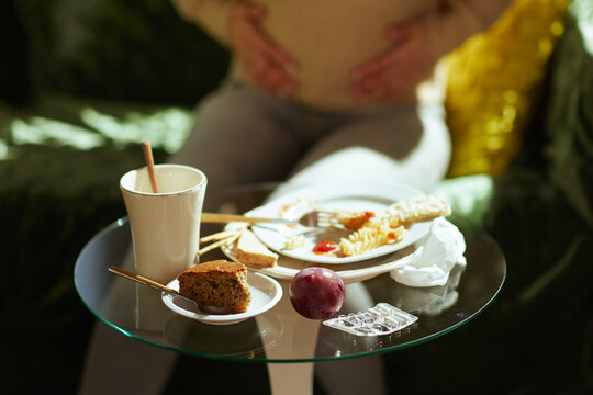 A person clutches their stomach in discomfort near a table littered with unhealthy food remains and medication, symbolizing the consequences of a poor diet.