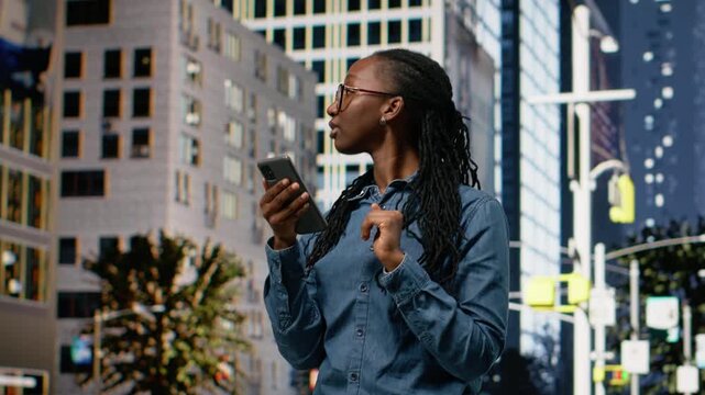 African American woman walking along city street using her smartphone, scrolling and browsing online content. Checking messages and following directions on navigation map app.