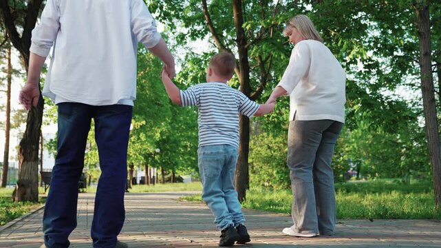 Family, kid boy playing holding parents hands, park summer. Family, dad mom, child son play together, happy child jumping. Family weekend, kid playing outdoors. Slow motion. Parents, child in nature