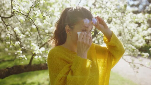 A woman wearing glasses lifts them to wipe her irritated eyes with a tissue, suffering from hay fever among blossoming trees in a sunlit park.