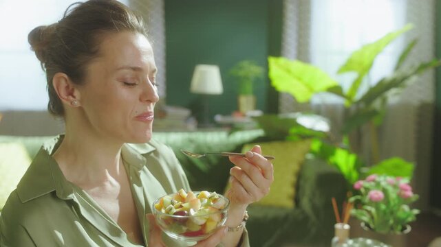 A happy woman enjoys a nutritious snack of mixed fruit salad in her living room, highlighting a healthy lifestyle and mindful eating habits.