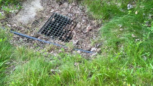 Park storm drain clogged with leaves and debris, aerial closeup of iron grate set in gravel and soil, dry leaf litter scattered