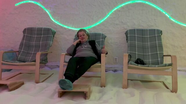 Elderly woman reclining in salt cave halotherapy chamber wearing cap on recliner under halogen neon lighting salt encrusted walls