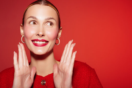 Smiling woman with red lipstick and hoop earrings poses with hands near face on red background. Concept of beauty, fashion, makeup, and confident female portrait.