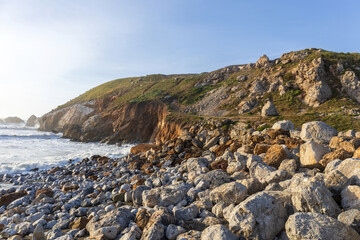 Fototapeta premium A rugged coastal landscape at Rockaway Beach in Pacifica, California, featuring a rocky shoreline scattered with large boulders and surf-worn stones beneath steep ocean cliffs.