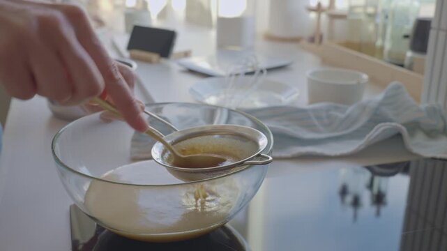 Hand strains creamy mixture through metal sieve into glass bowl in kitchen. Concept of baking preparation and home cooking