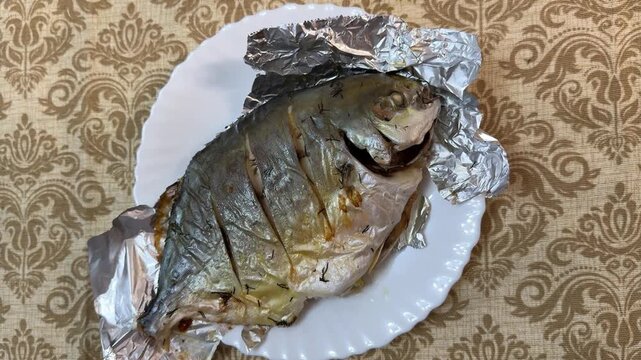 Golden pompano grilled on foil plate, overhead view showing two cooked fillets resting on crinkled aluminum foil and white ceramic