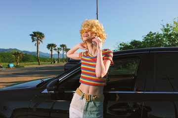 Fototapeta premium Smiling woman with curly blonde hair leaning beside a black car in a sunny outdoor scene, wearing a rainbow striped top and light shorts, conveying carefree retro summer lifestyle.