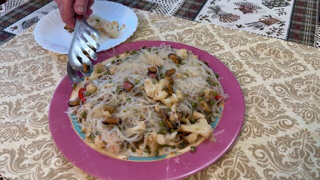 Female hand serving noodles and mussels on porcelain plate, tongs portioning funchoza with cauliflower, peas, red pepper and oil
