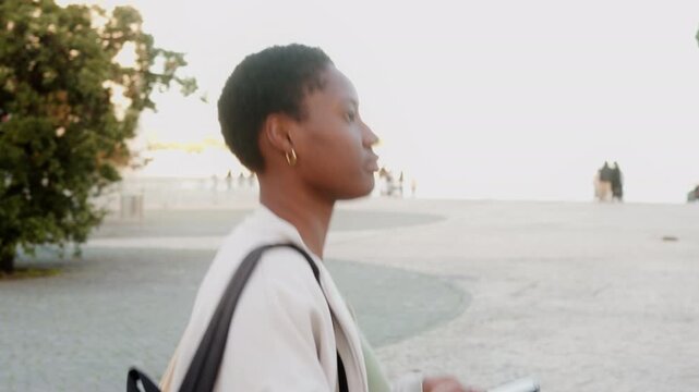 Young Black woman walking while using smartphone, focused on screen as she moves along bright open walkway carrying backpack in casual during daytime