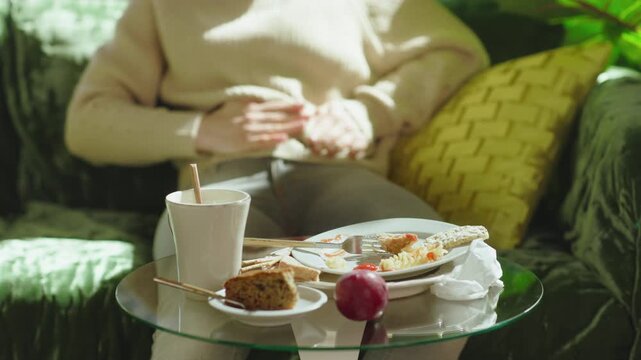 A woman in a beige sweater rubs her abdomen in a circular motion on a sofa, experiencing bloating and indigestion after eating a heavy meal at home.