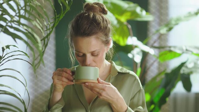 A happy woman sips from a green ceramic mug, enjoying a quiet moment of contentment and relaxation among her indoor plants in a sunlit room.
