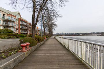 Fototapeta premium Quiet Waterfront Boardwalk Along River With Residential Buildings, Trees, and Railings in New Westminster