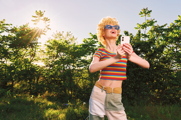 Fototapeta premium Cheerful woman outdoors in retro style, striped crop top and sunglasses, golden sunlight, relaxed pose, capturing moment on phone, lush trees in background, warm natural tones against a clear sky