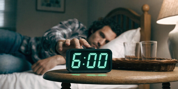 Tired man waking up in bed reaching for a digital alarm clock showing 600 AM