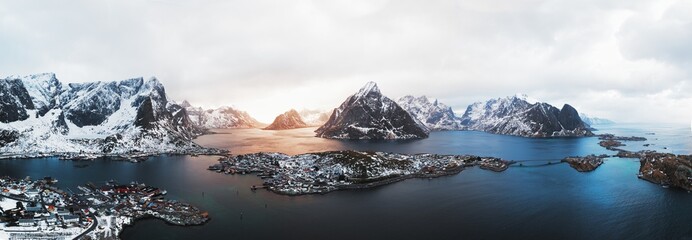 Atmospheric drone panorama of Reine and nearby islands in the Lofoten archipelago, northern Norway. Golden hour light reflects across Arctic fjords between snowy mountains and remote fishing villages. © Eclipsnet