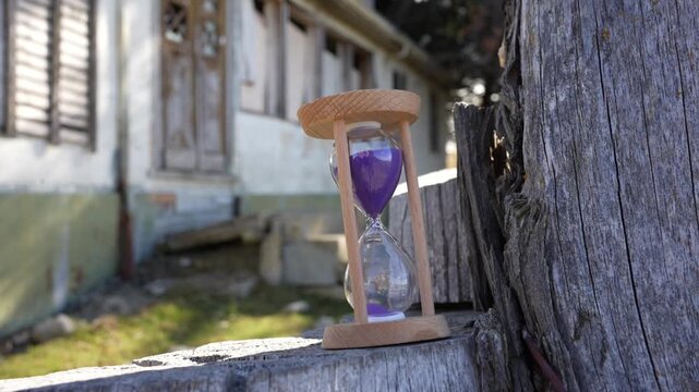 Close up of sand timer running out with abandoned vintage house background, Wooden hourglass with purple sand on old fence post