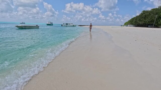 Woman in bikini walks along sandy beach with boats in Fulidhoo local island in Maldives, ocean waves gently lapping at her feet.
