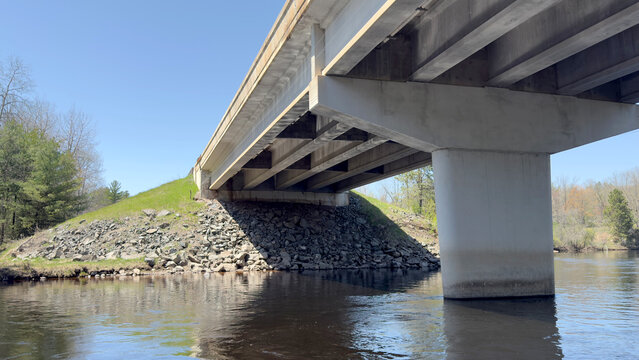 Low-angle view of a modern concrete highway bridge crossing a calm river, supported by a large central pillar with riprap stone embankment.