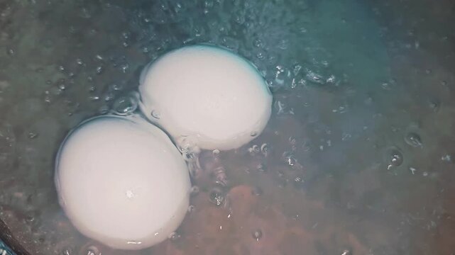 Two white chicken eggs boiling in a pot of hot water with bubbles, close-up top view of home cooking preparation.