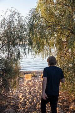 Man walking to two empty camping chairs sitting on a sandy water front as the sun sets across Lake Pamamaroo, Menidee Lakes, New South Wales, Australia