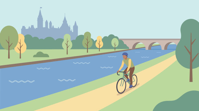 A cyclist rides along a river path with a cityscape in the background near a bridge and trees
