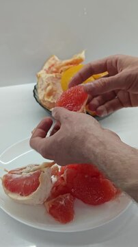 A detailed vertical shot captures a mans hands meticulously removing the peel and white pith from a ripe red grapefruit, separating the juicy segments onto a pristine white plate, highlighting a