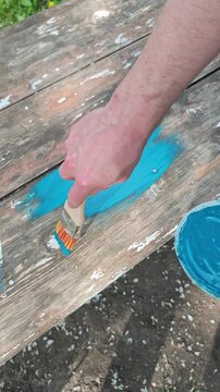 Detailed shot of male hands actively painting an aged and distressed wooden plank with bright turquoise paint using a paintbrush, highlighting home improvement, DIY repair, and outdoor furniture