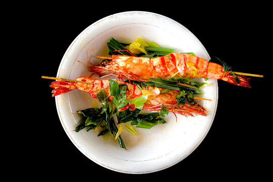 Acehnese style fried prawns on skewers with aromatic curry leaves in a white bowl, top view isolated on black background.