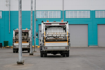 Naklejka premium Two white garbage trucks are parked in a lot outside an industrial building. The building features teal and white walls with large grey doors on a daytime