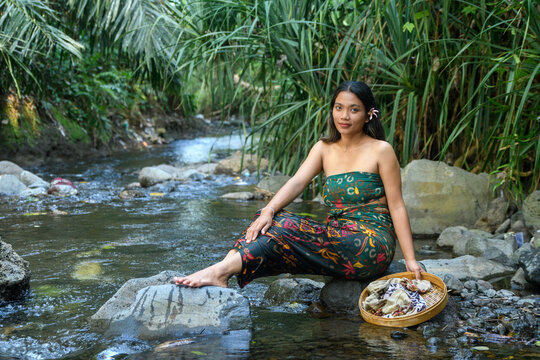 Balinese woman in dark green floral strapless dress sitting on river rock splashing water with woven basket