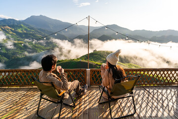 Naklejka premium Young couple relaxing and enjoying the sunrise and sipping their morning coffee amidst the beautiful mountain and misty scenery
