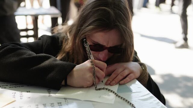 Close portrait of woman with sunglasses writing on ema plaque with chained pen at outdoor shrine Japan