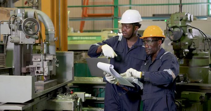 two african adult male engineers coordinate near milling machine, one points while one checks document in workshop, safety teamwork, production planning and equipment review