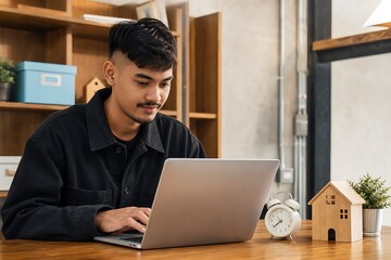 Fototapeta premium Focused young man using a laptop at a desk with a small plant and clock nearby