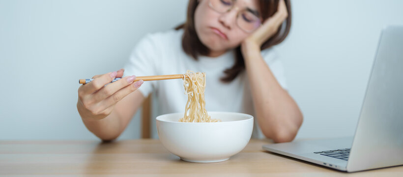 Bored young Asian woman working late at night on laptop while a bowl of hot instant ramen noodles at home. Unhappy female having a quick meal during work from home. Simple lifestyle concept