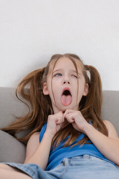 Young playful girl with pigtail making silly expression face tongue out feeling bored sitting on grey couch wearing blue shirt white background casual childhood home environment