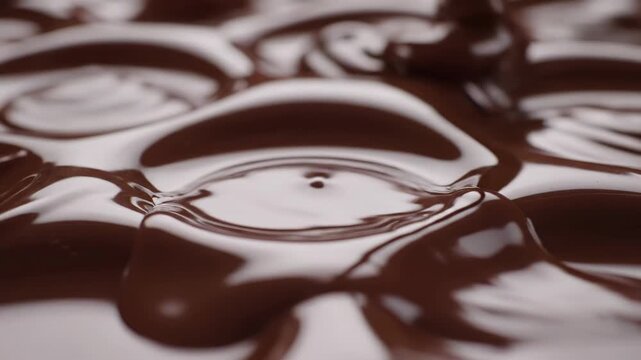 Close-up macro shot of melted milk chocolate being poured, creating circular ripple patterns and glossy reflections on a smooth viscous surface with soft cinematic studio lighting.