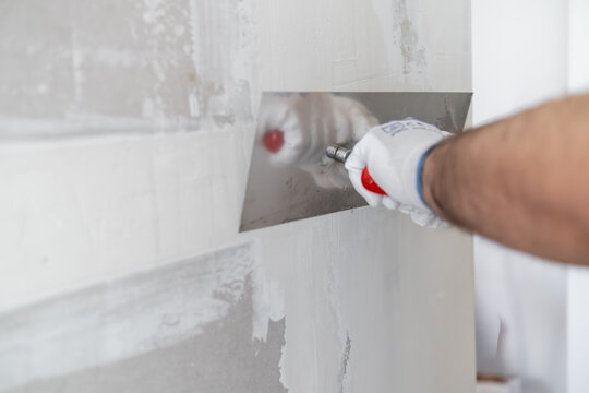 Professional applying a smooth finishing coat to drywall using a stainless steel trowel. Clean and bright working environment with prepared walls visible in the background.