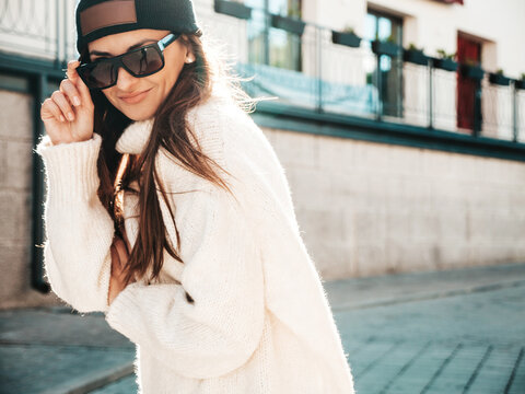 Portrait of beautiful smiling model. Female dressed in warm hipster white sweater and beanie. Trendy posing in the street at sunset. Funny and positive woman having fun outdoors in sunglasses