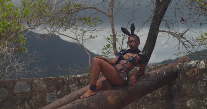 A dark-skinned woman in a luxurious bunny outfit posing atop the weathered stone walls of a Caribbean fort with crashing waves below.