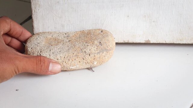 Close up of Asian man placing a stone as a simple door stopper on tiled floor near wooden door indoors, preventing door movement with improvised household object.