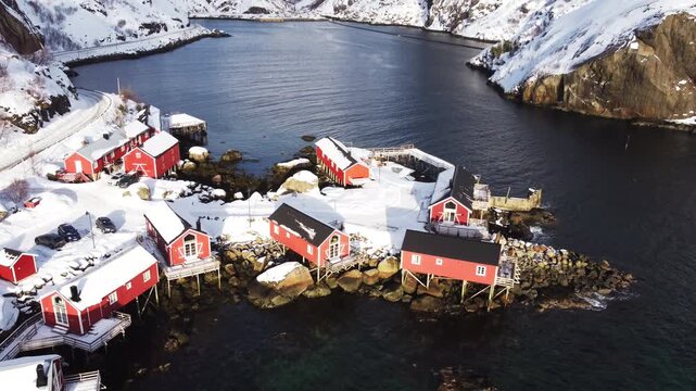 Drone shot over red rorbuer in Nusfjord historic fishing village with snowy mountains background, Lofoten Islands, Norway. Top down perspective of iconic arctic coastal architecture