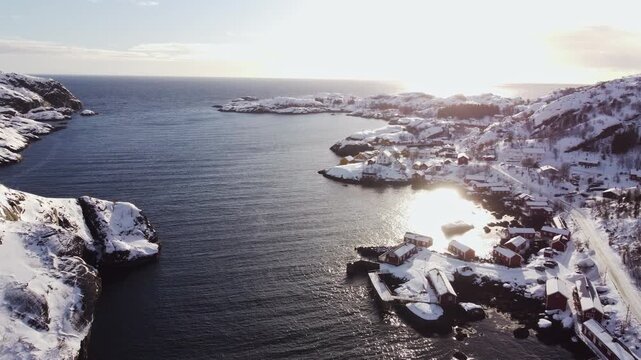 Aerial drone perspective of a snowy coastal town in Lofoten Islands with sun shining through quiet ocean waters. Calm arctic winter morning scene in Northern Norway archipelago