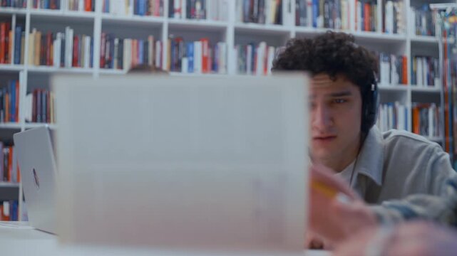 A focused university duo studies together in a quiet library. The student on the left watches an online lecture through headphones, and his partner reviews handwritten workbook pages.
