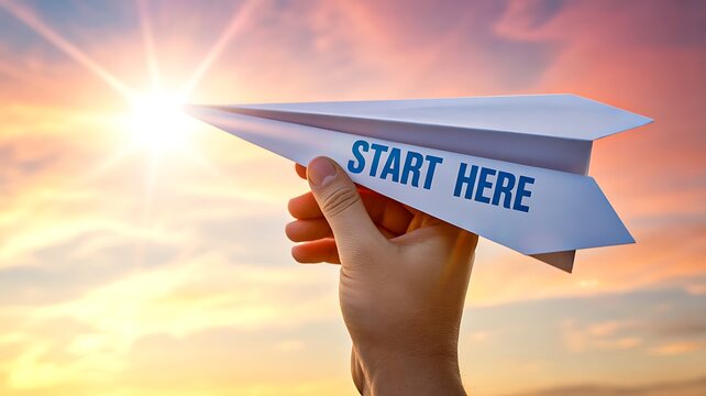 Hand holding paper airplane with start here message at sunset sky