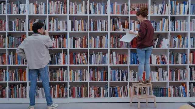 Two young people looking for specific books on a large white bookshelf in a public library