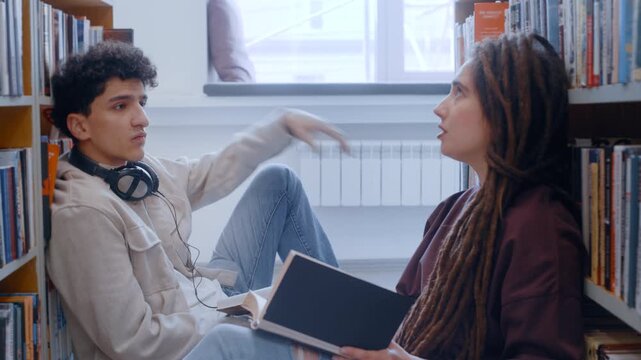 Two young learners sit tucked between library shelves, completely lost in conversation with open books resting on their knees. Their animated discussion brings the stories and theories to life