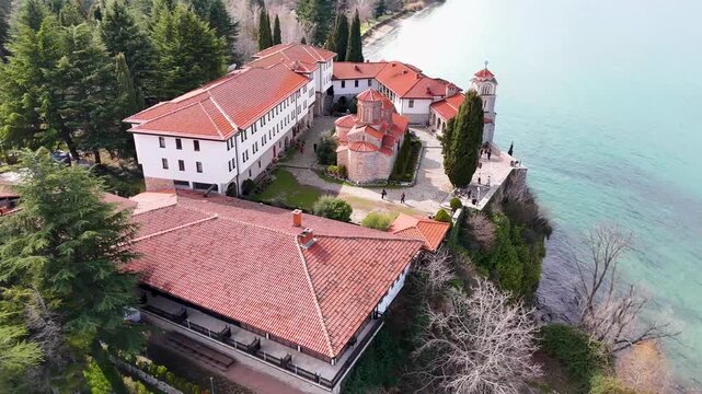 Aerial drone tracking shot orbiting counterclockwise around the historic Eastern Orthodox Saint Naum Monastery by the crystal waters of Lake Ohrid