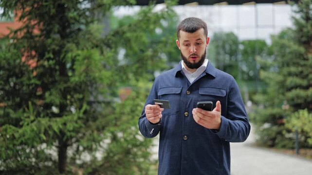 Young bearded man surprised by missing money while checking phone and credit card outdoors. Person faces bank account theft, unexpected fees or financial fraud. Concept of cyber crime and stress.